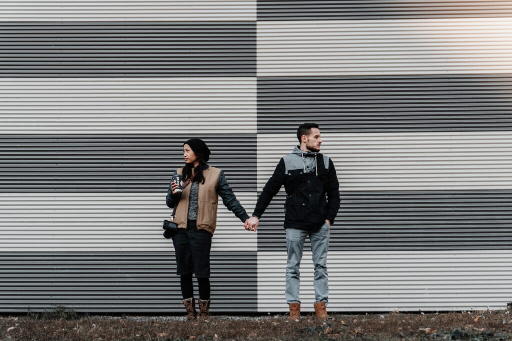 Man and woman standing infront of a striped metal wall, holding hands but looking in opposite directions