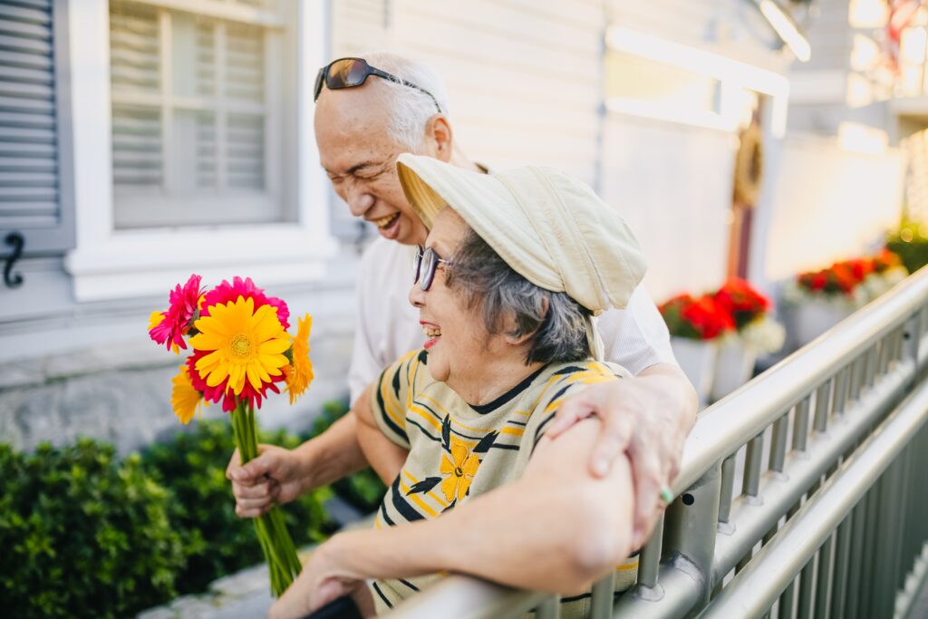 Elderly couple standing along a railing. The man holds flowers in his hand and both are laughing