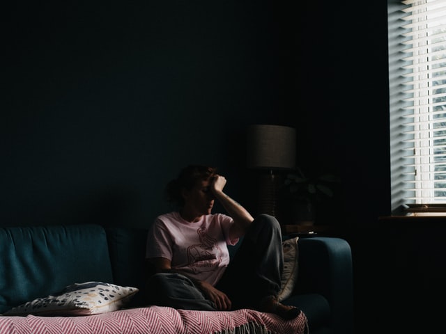 Somber image with a person sitting on couch, looking sad with their head resting against their hand.