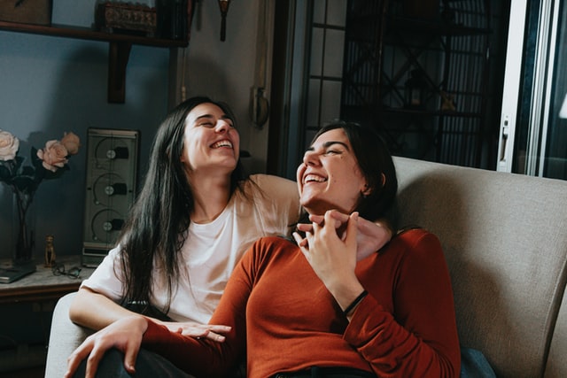 Female couple leaning on each other, smiling with eyes closed and intertwined fingers on a couch.