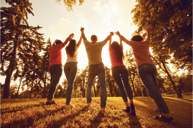 A group of five young people holding hands as they raise them standing in a forest clearing